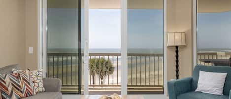 A living room with a sofa, a blue armchair, a wooden coffee table, and a view of the beach through a sliding glass door.