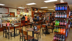 Un restaurante con mostrador de deli, mesas, sillas y estante para bebidas.