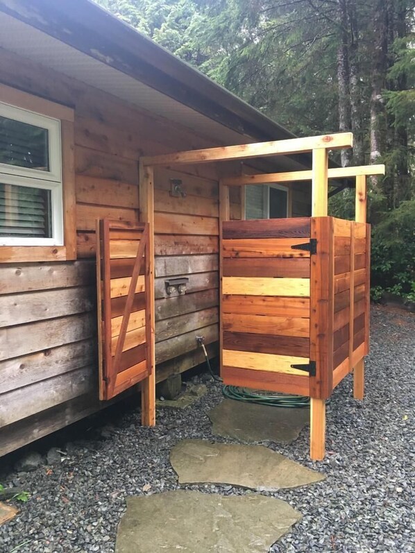 Una cabaña de madera con la puerta abierta, mostrando una ventana y un pequeño espacio exterior con grava y piedras para pisar.