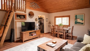 A spacious living room with a wooden ceiling, a TV, a dining area with wooden chairs, and a staircase.