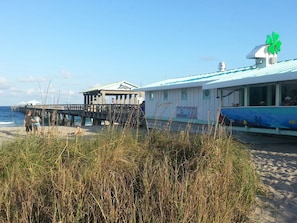 Un edificio frente a la playa con un muelle que se adentra en el mar, una zona techada y un cartel que promociona la adopción de tortugas marinas.