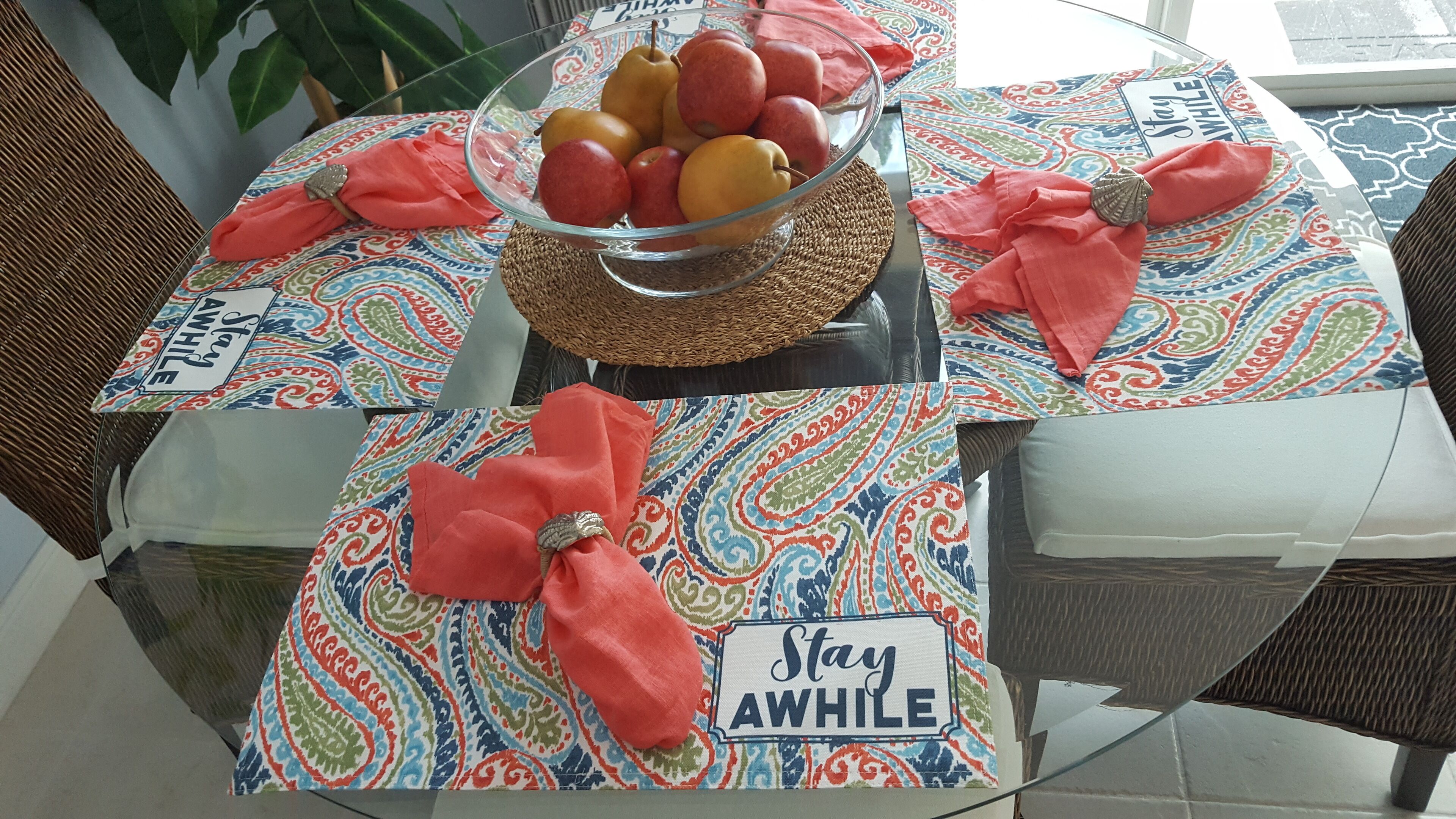 Una mesa con cubierta de vidrio, un cuenco de fruta, un mantel individual con el texto 'Stay Awhile' y un mantel estampado.