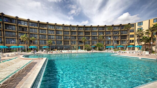Ein Hotel mit einem großen Pool, umgeben von Liegestühlen und Sonnenschirmen, ein mehrstöckiges Gebäude mit Balkonen und Palmen.