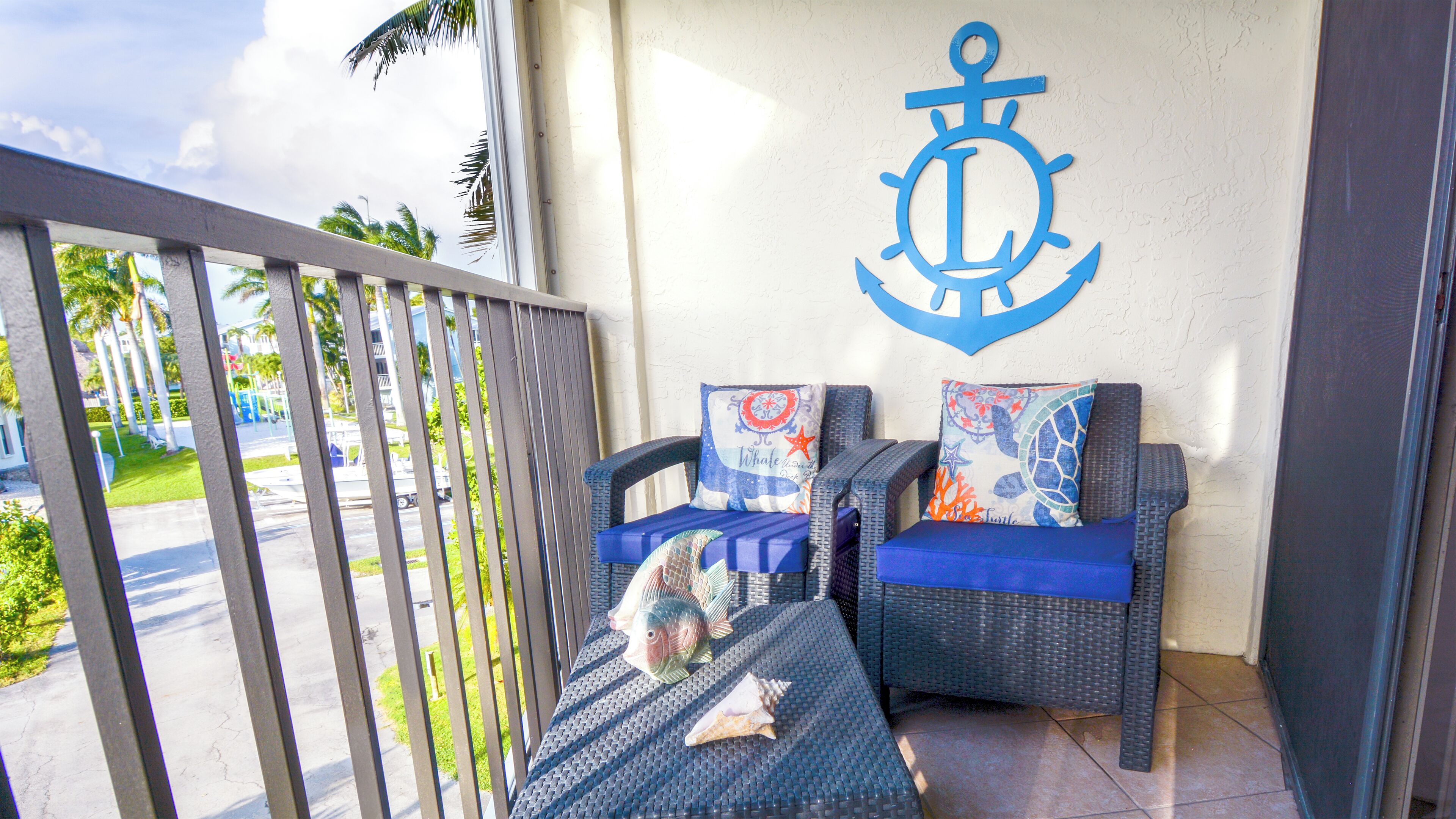 A balcony with wicker chairs, a wall with an anchor decoration, and a view of palm trees and a street.
