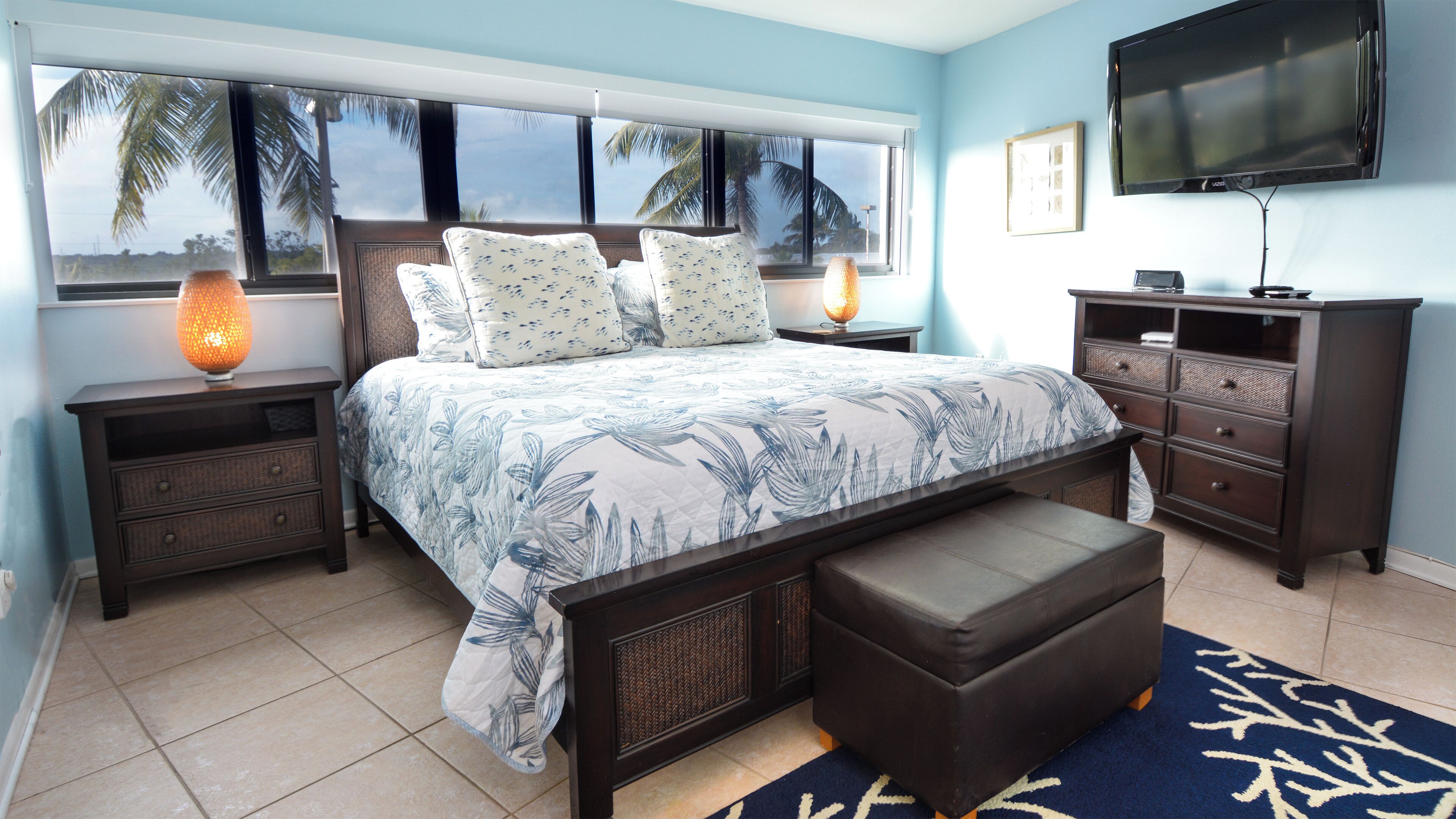 A bedroom with a bed, nightstands, a dresser, a TV, and a view of palm trees through large windows.
