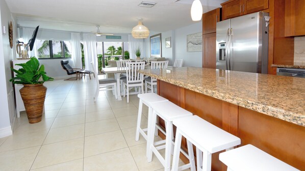 A modern kitchen with a granite countertop, white bar stools, and a stainless steel refrigerator.