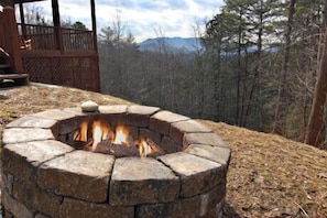 Ein Steinkamin mit Feuer, umgeben von einer Holzterrasse und einer bewaldeten Landschaft mit Bergen im Hintergrund.