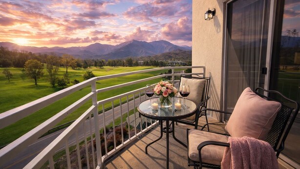 A balcony with a table set for two, a chair, a cushion, and a view of a golf course and mountains.