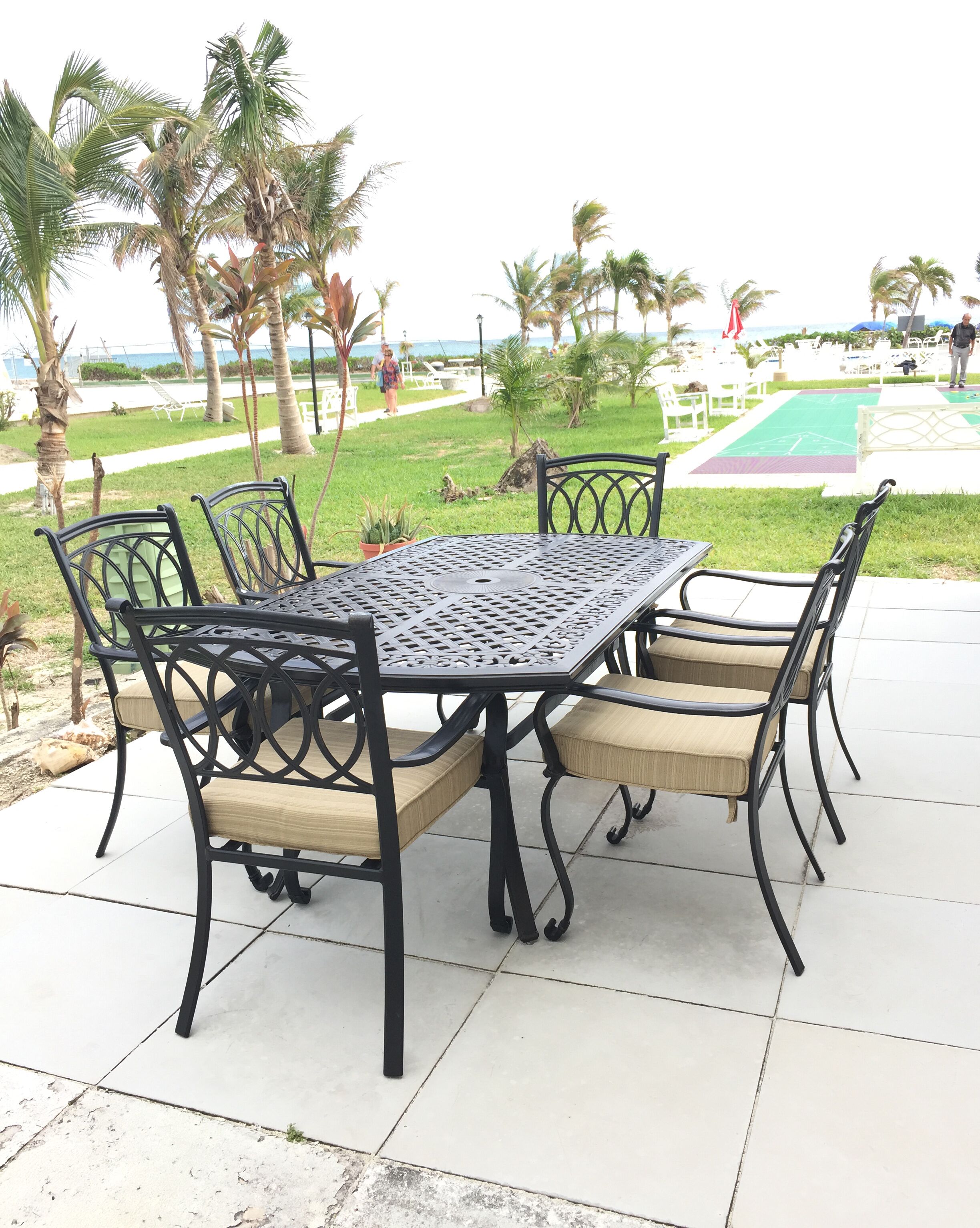 An outdoor patio with a metal table and chairs, surrounded by palm trees and a view of the beach.