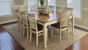 A dining room with a wooden table and six chairs, surrounded by beige walls and large windows with blinds. -  Make Memories at Barefoot Daze (Kitty Hawk)