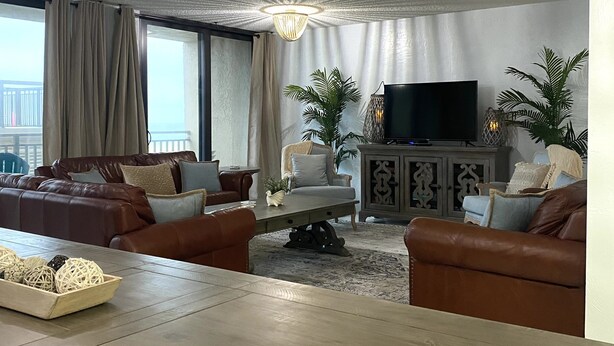 A modern living room with a large wooden table, leather sofas, a television, and decorative plants.