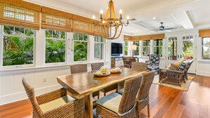 A spacious dining area with a wooden table and wicker chairs, large windows with tropical views, and a chandelier. - Hale Wai'olu by Alekona Kauai: Luxury 3br ocean view home in Kukuiula, AC, private pool, fire pit (Koloa)