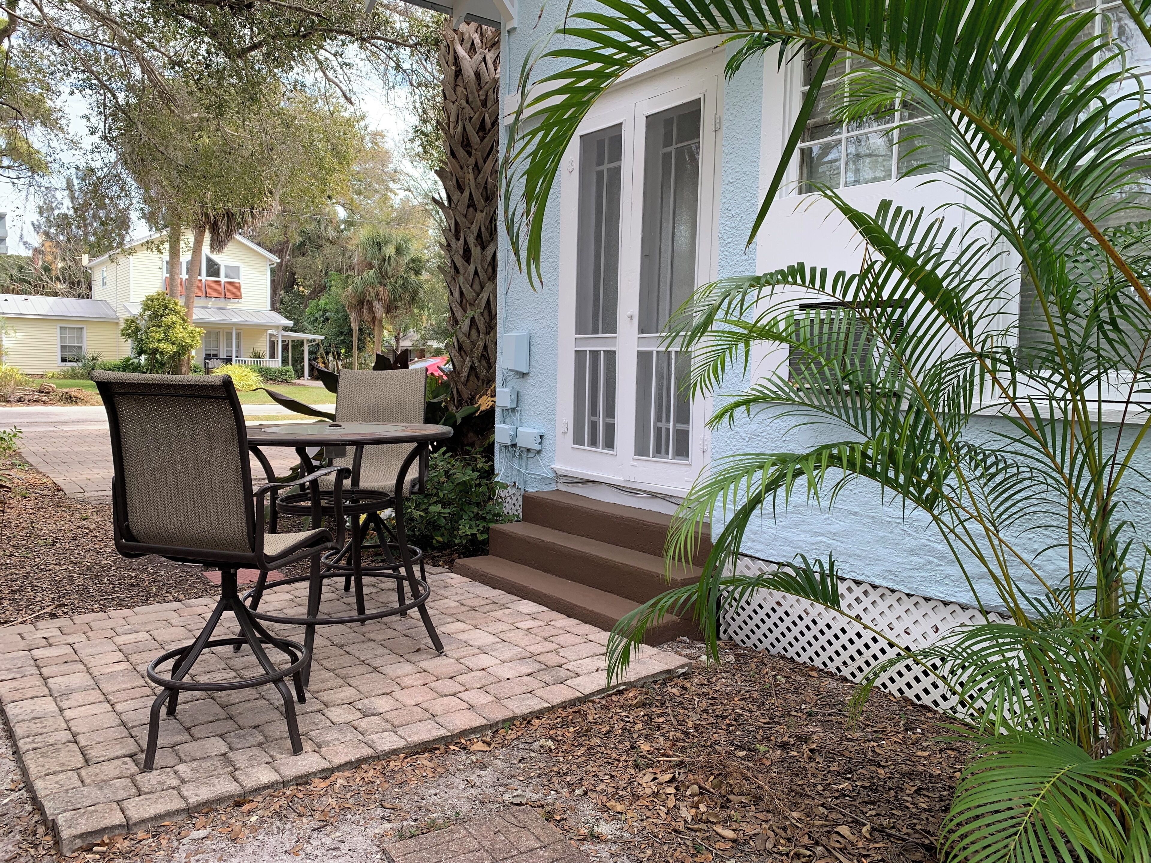 Eine Terrasse mit Tisch und Stühlen, eine weiße Tür und ein kleiner Gartenbereich mit einer Palme.