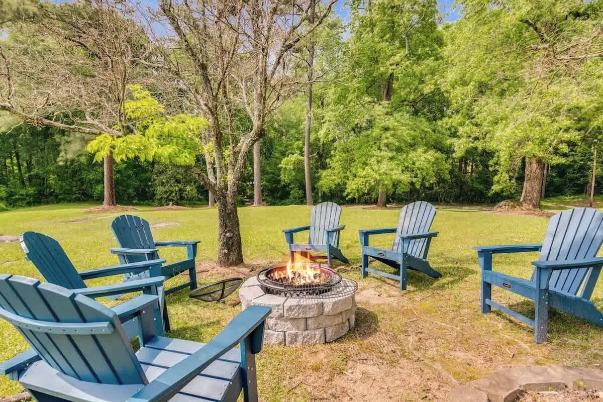 A fire pit surrounded by Adirondack chairs in a grassy outdoor area with trees.