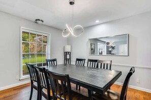 Zona de menjador amb taula rectangular, sis cadires, lampadari modern, mirall gran i finestra amb persianes.