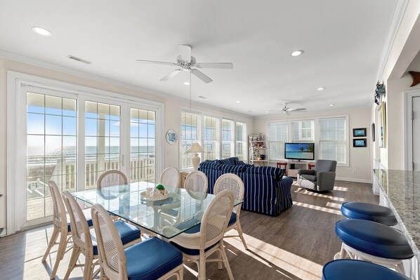 A spacious living area with a glass dining table, white wicker chairs, a striped sofa, and a TV.