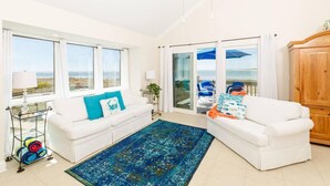 Un salon lumineux avec deux canapés blancs, un tapis à motifs bleus et une vue sur la mer grâce à de grandes fenêtres.
