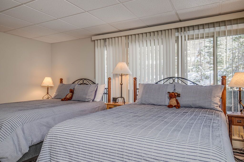 A hotel room with two beds, each with striped bedding and a teddy bear on the pillows. There are bedside lamps and a window with blinds.