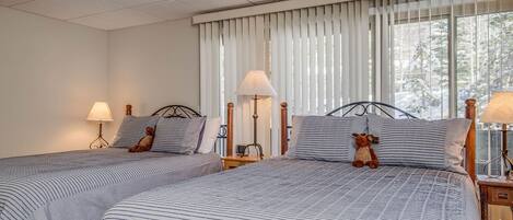 A hotel room with two beds, each with striped bedding and a teddy bear on the pillows. There are bedside lamps and a window with blinds.