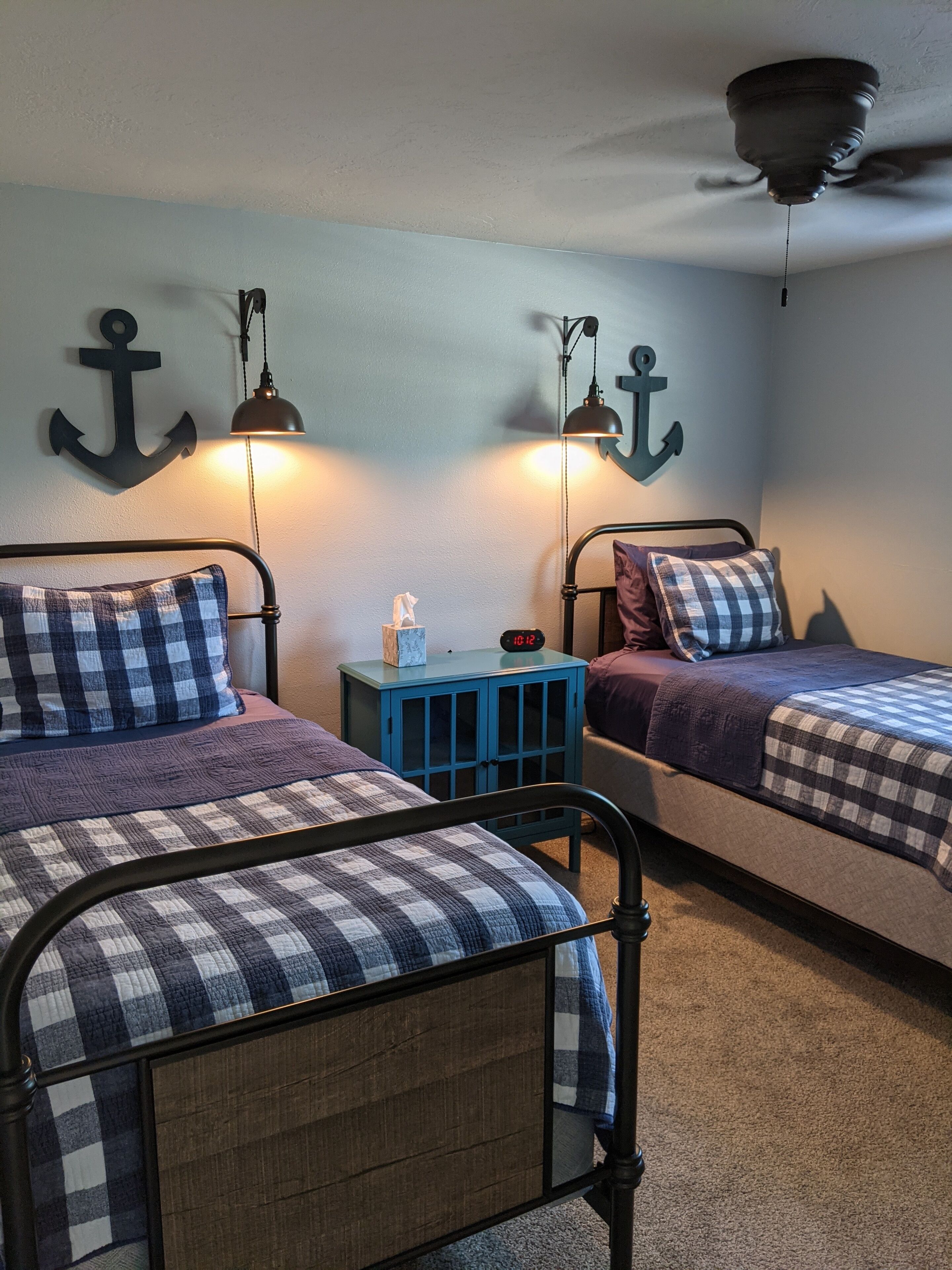A bedroom with two beds, each with a checkered blanket and pillows, a small blue nightstand with a clock, and wall-mounted lamps.