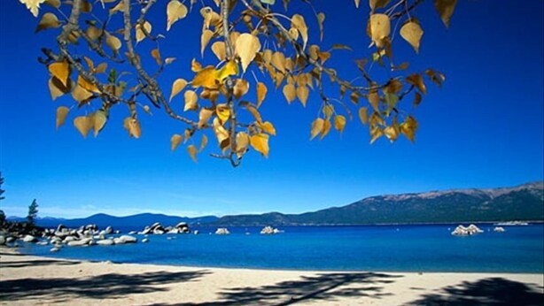Une plage avec une eau d’un bleu limpide, un rivage rocheux et des montagnes en arrière-plan.