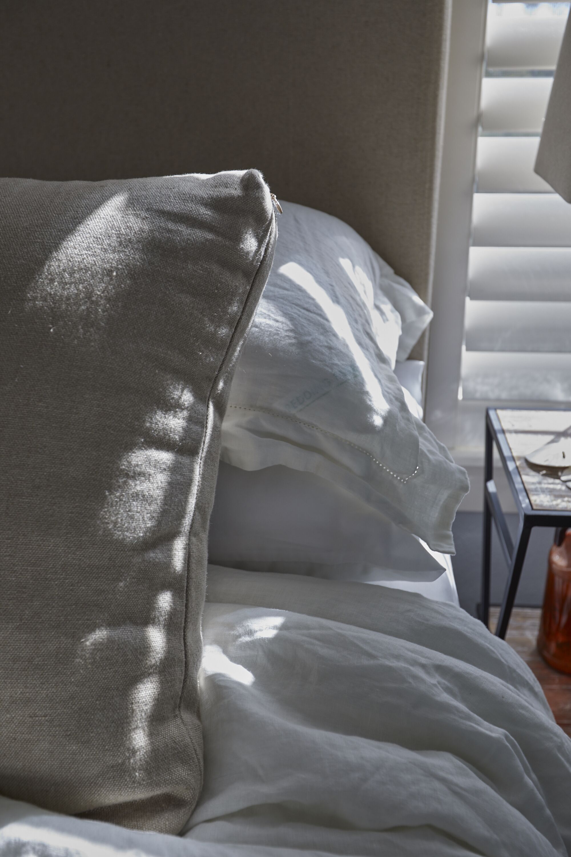 A bed with white bedding and a grey pillow, a small table with a book, and a window with blinds.
