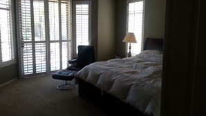 A bedroom with a large bed, a chair, and a lamp. There are windows with blinds and a view outside. - Estrella's NASCAR HOT SPOT! FALL GOLF GETAWAY! HOLIDAY PLAY! (Goodyear)
