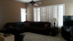A living room with a sectional sofa, a television, a ceiling fan, and a large window with blinds. - Estrella's NASCAR HOT SPOT! FALL GOLF GETAWAY! HOLIDAY PLAY! (Goodyear)