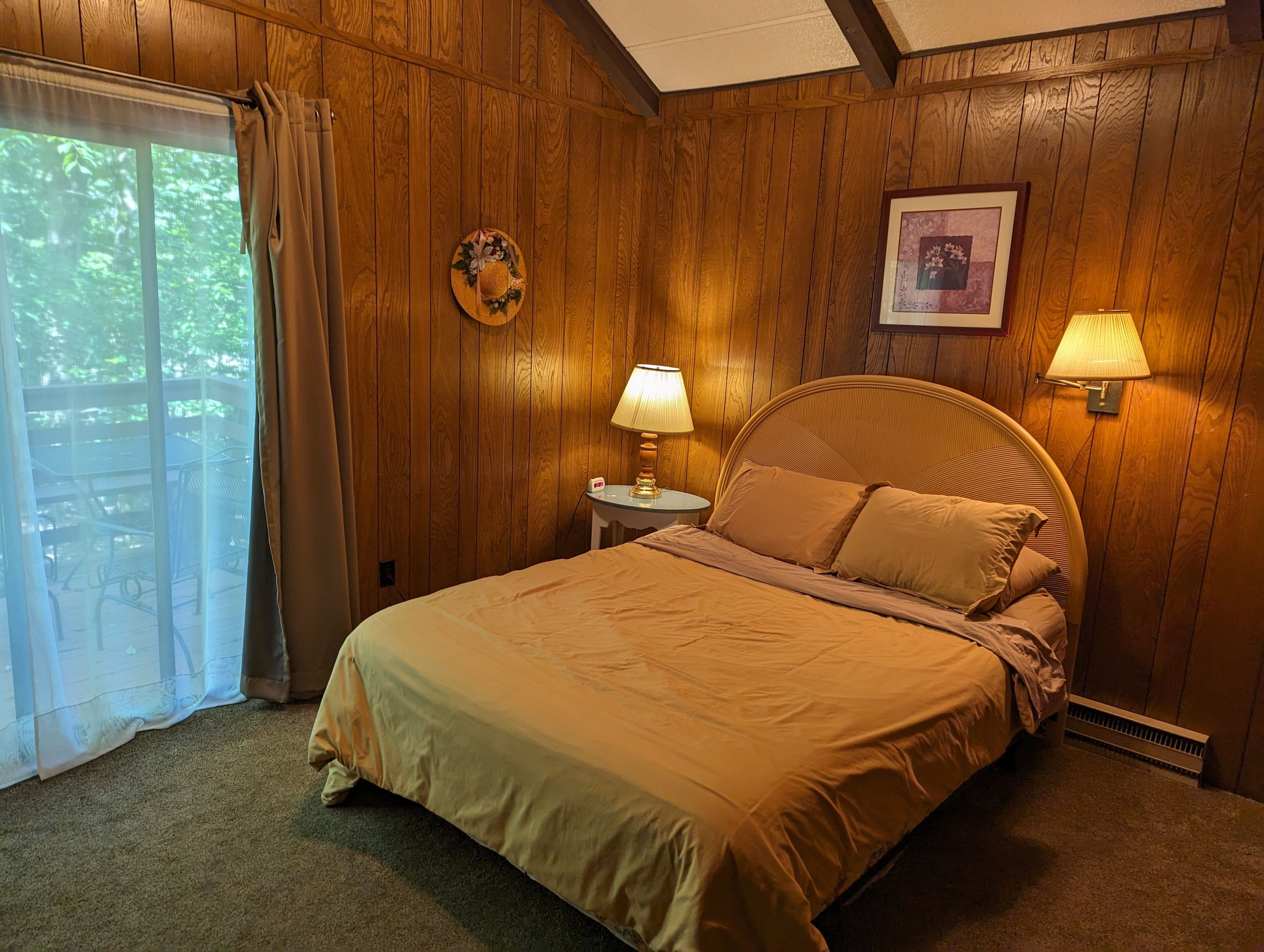 Une chambre avec un mur lambrissé en bois, un lit avec une tête de lit, deux lampes de chevet, un tableau encadré et une fenêtre avec des rideaux.