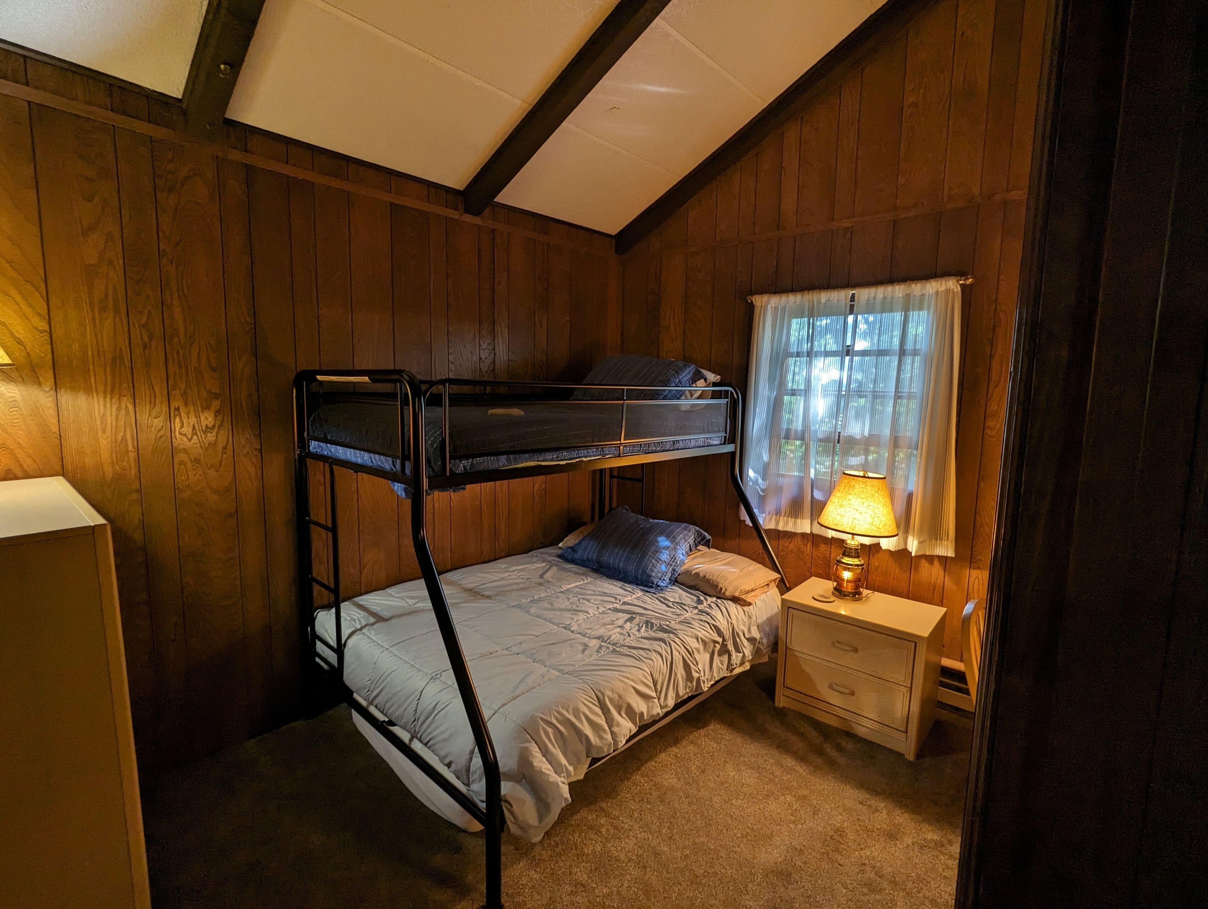 A bunk bed room with a window, a bedside table, and a lamp.