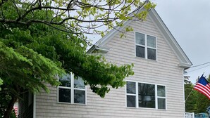Une maison à deux étages, avec une façade blanche et un jardin devant. On y trouve deux chaises Adirondack en bois et une petite table.