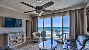 A spacious living room with a large window offering a view of the beach, a ceiling fan, a flat-screen TV, a leather sofa, and a glass coffee table. - 310 Sterling Sands Wake Up to Ocean Views in This Luxurious Beachfront Oasis! (Destin)