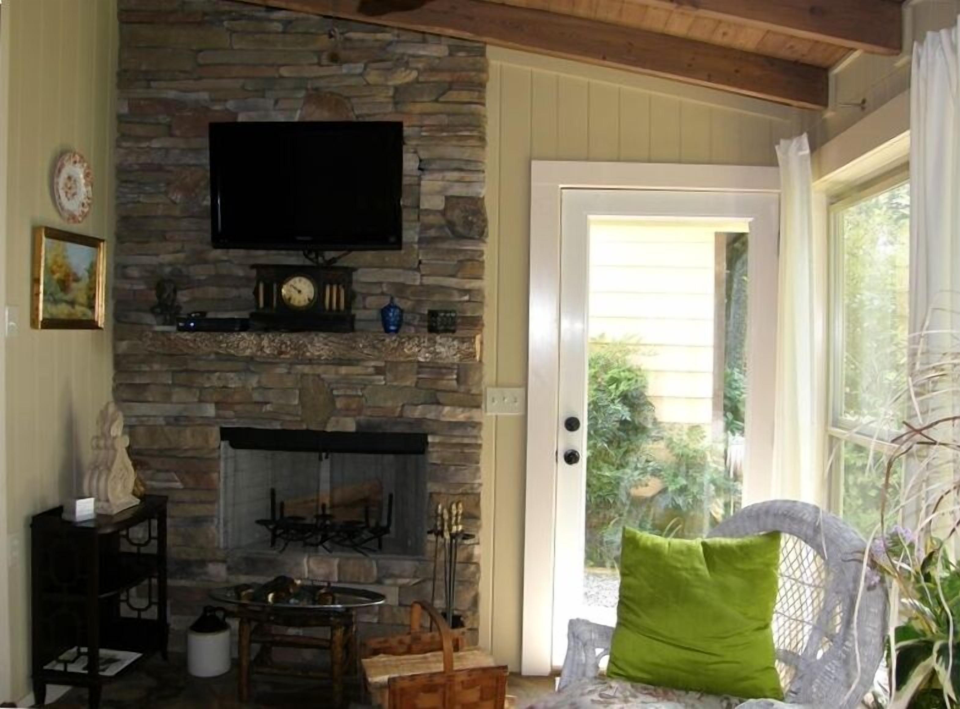 A cozy room with a stone fireplace, a flat-screen TV mounted above it, and a door leading to a garden.
