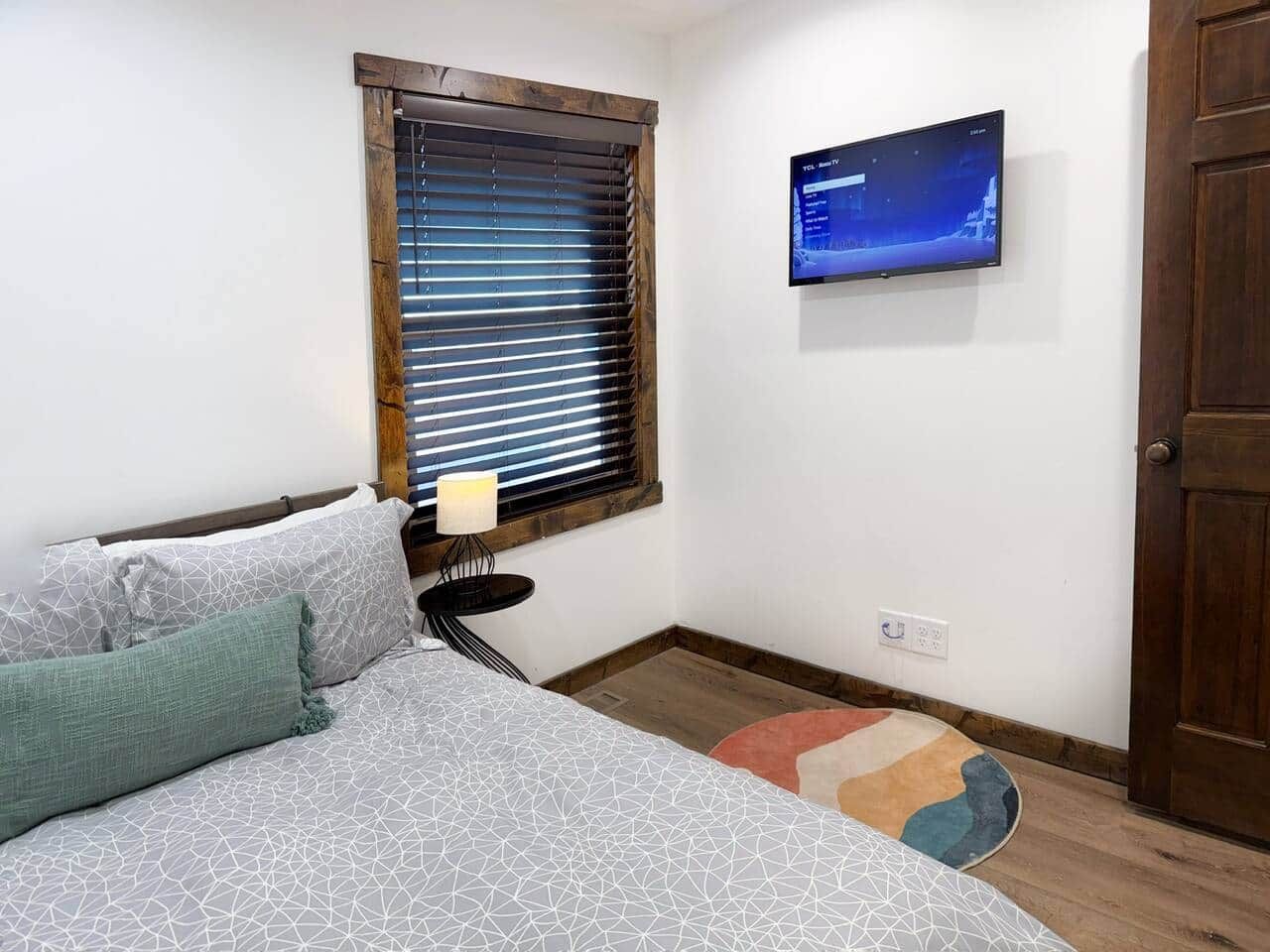A bedroom with a bed, a TV mounted on the wall, a window with blinds, and a door.