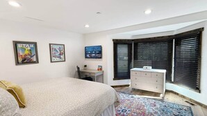 Ein Schlafzimmer mit Bett, Schreibtisch, Kommode und Fenster mit Jalousien.