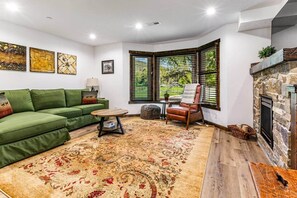 Un salon avec un canapé vert, une table basse ronde, un fauteuil en cuir, une cheminée et une grande fenêtre avec des stores.