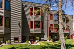 Un immeuble à plusieurs étages, à la façade grise et aux encadrements de fenêtres rouges, doté de balcons et d’une pelouse bien entretenue.