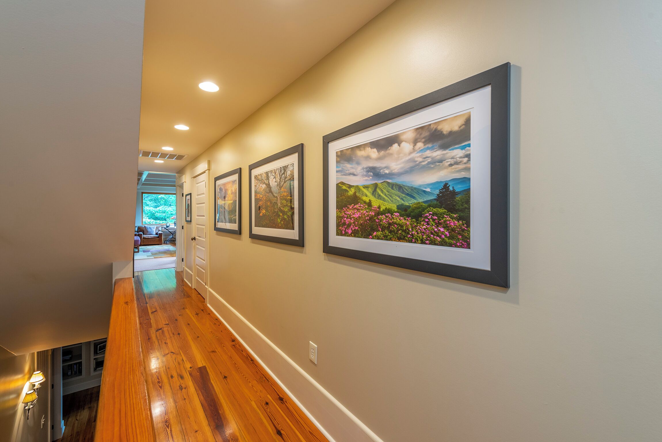 A hallway with wooden flooring, recessed lighting, and framed landscape pictures on the wall.