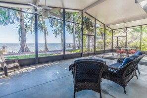 Un patio techado con muebles de mimbre, ventanas amplias que ofrecen vistas al agua y a los árboles.