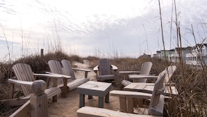 Een strandsetting met houten stoelen en tafels op het zand, omgeven door hoge grassen en enkele huizen op de achtergrond.