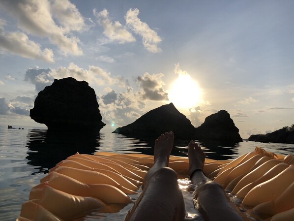 A person reclining on an inflatable raft with a view of the sun setting behind two large rock formations.