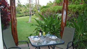 An outdoor dining area with a table set for two, surrounded by chairs, in a garden setting with various plants and trees. - Fairways 1706 - Renovated, includes Beach Access, Bikes (Kamuela)