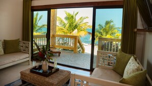 Habitación con sofá, una mesa de centro de mimbre y vista a la playa y palmeras a través de puertas de cristal grandes.