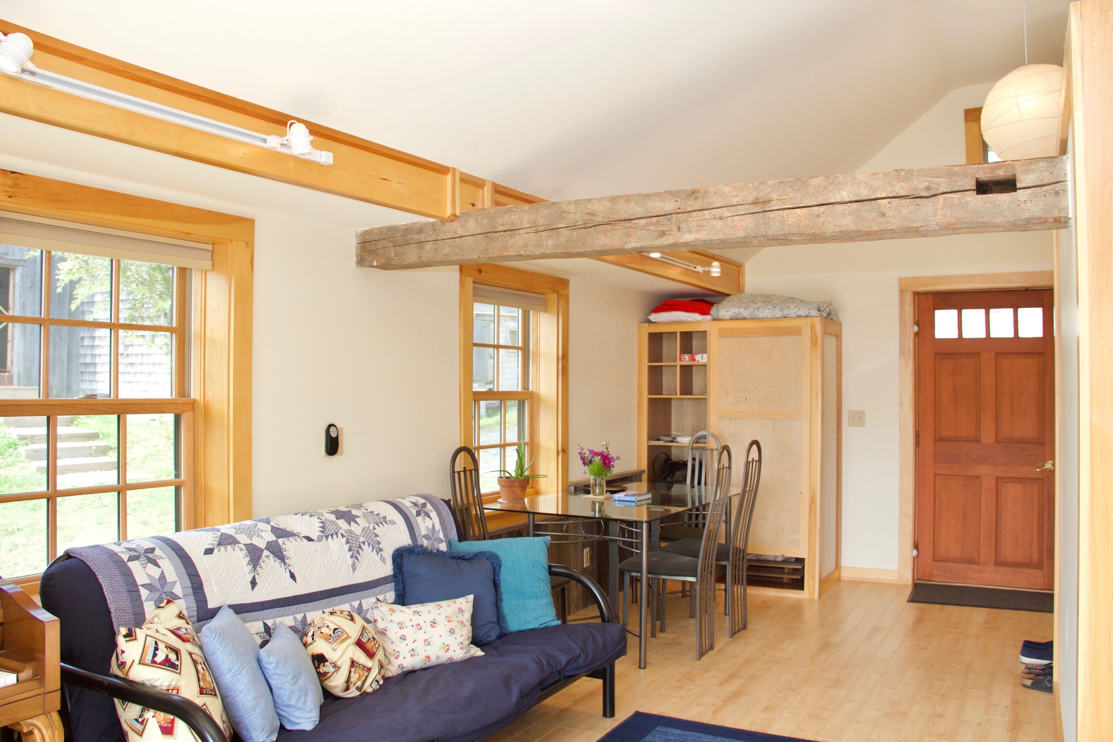A cozy living room with a sofa, dining table, and chairs. There is a wooden ceiling beam, a door, and a window.