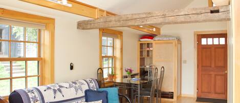 A cozy living room with a sofa, dining table, and chairs. There is a wooden ceiling beam, a door, and a window.