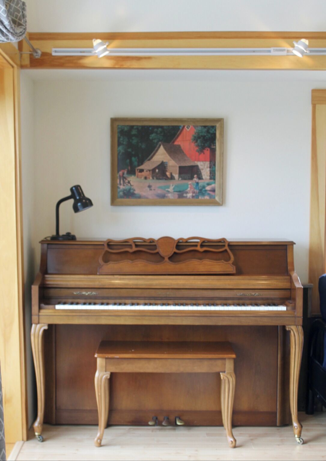 A wooden piano with a bench in front, a painting on the wall, and a lamp to the left.