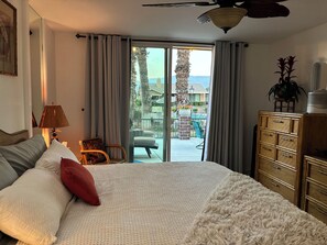 A bedroom with a large bed, a wooden dresser, a lamp, and a view of a pool and outdoor seating area.