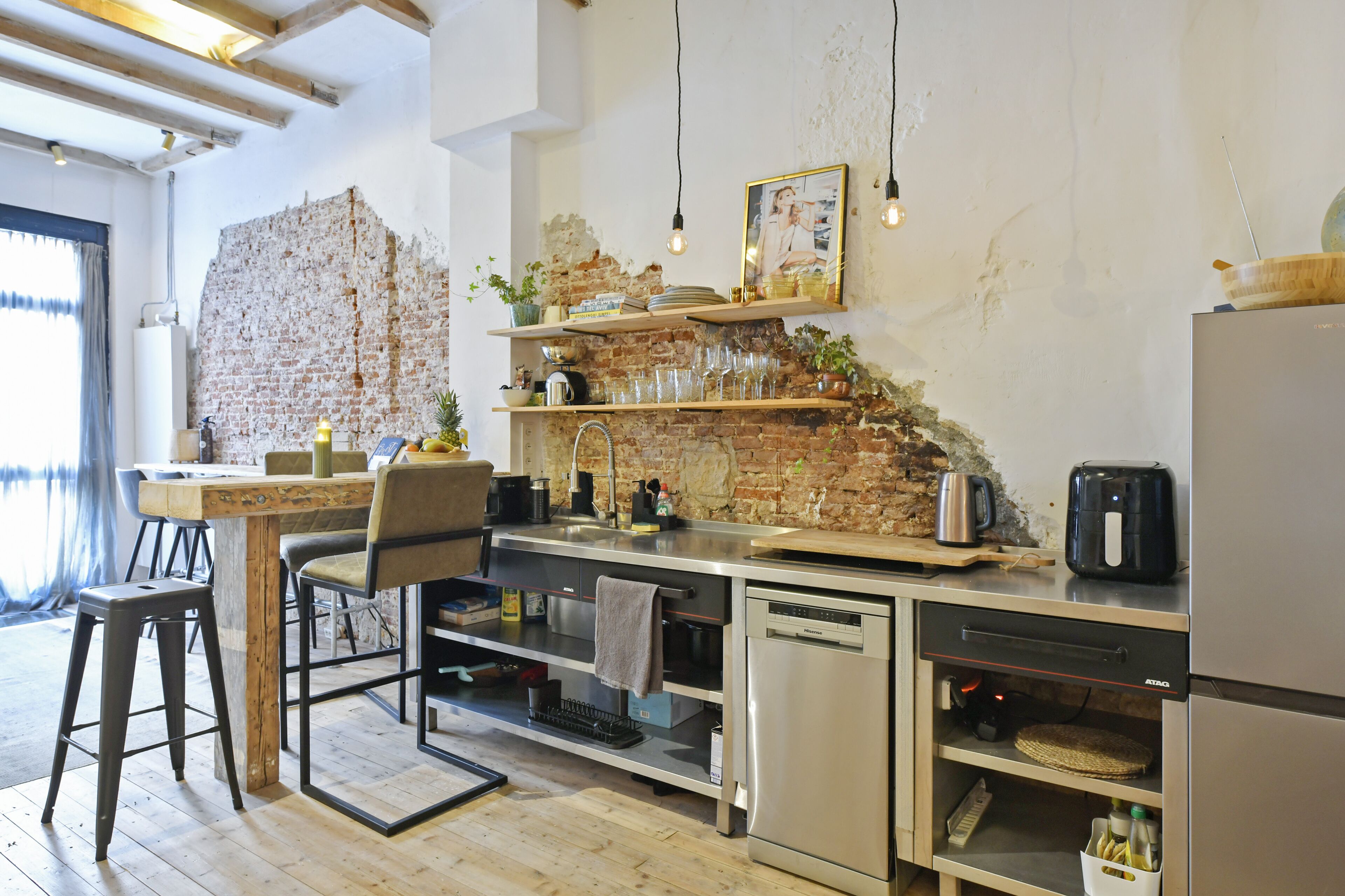 Une cuisine moderne avec des murs en briques apparentes, un plancher en bois et des appareils électroménagers en acier inoxydable. On y trouve des tabourets de bar, une machine à café et divers ustensiles de cuisine.