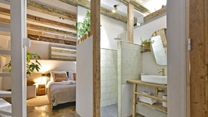 Baño con tocador de madera, lavabo y espejo. Hay una zona de ducha con puerta de cristal y una planta pequeña sobre el tocador.
