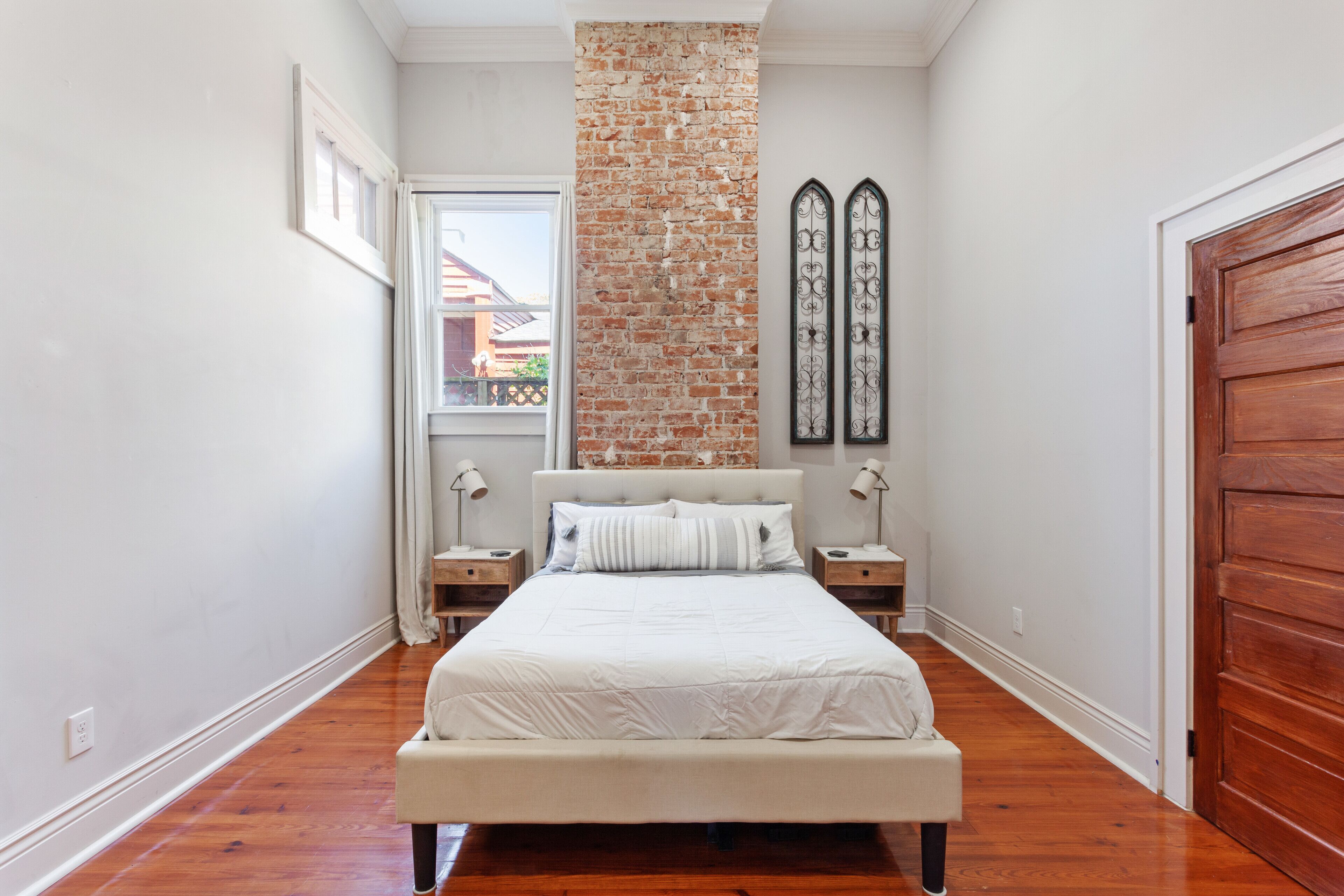 A bedroom with a brick accent wall, a large bed with white bedding, two bedside tables with lamps, and a wooden door.